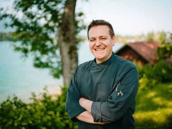 Lächelnder Koch in dunkler Uniform steht im Freien nahe einem See und einer Hütte